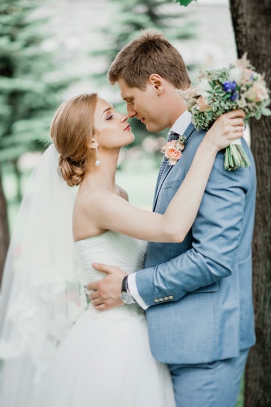 Groom in a pale blue suit kissing the bride among pine trees